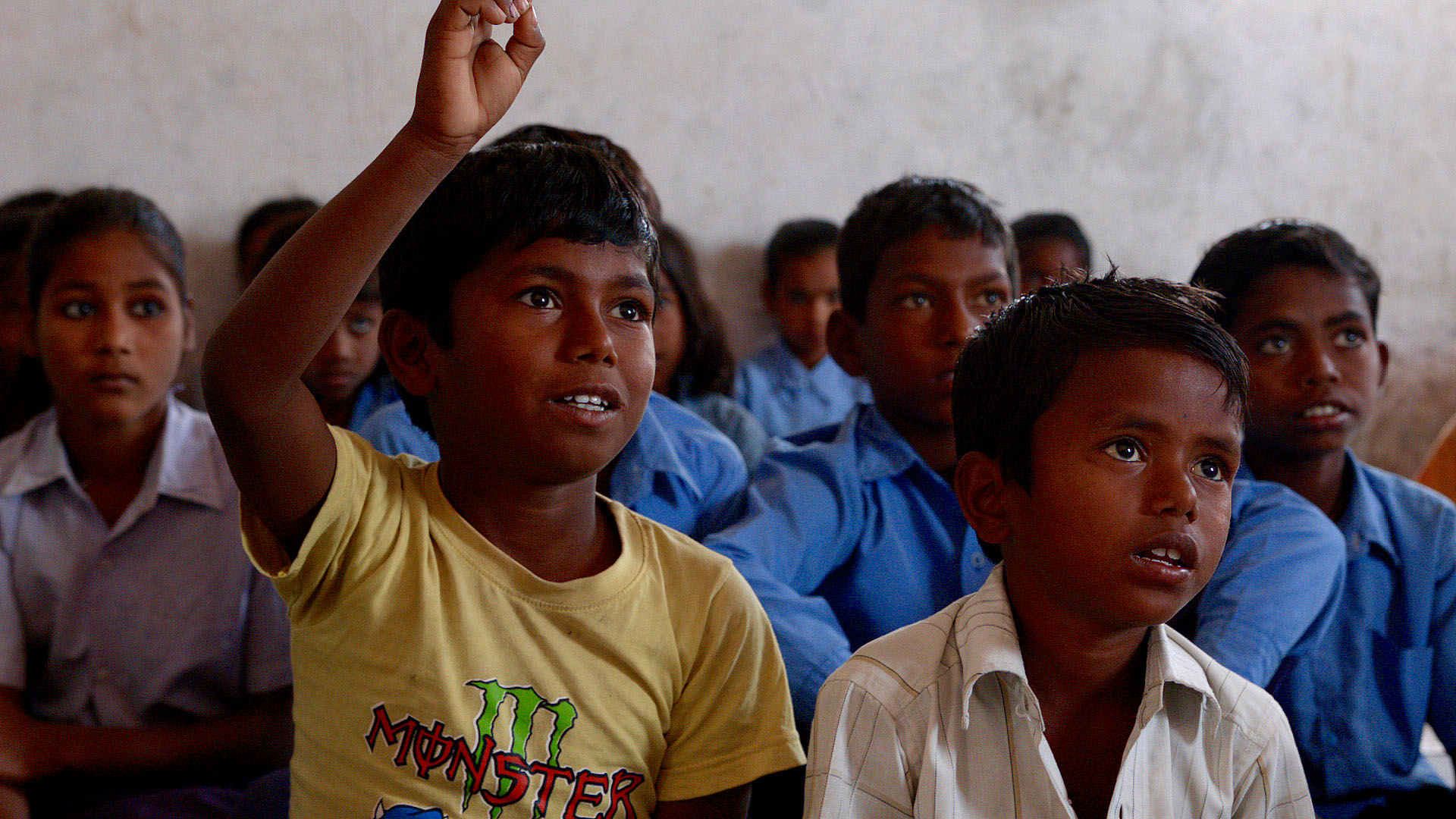 Children sit in a classroom, focused on something off-camera. One boy in the front row raises his hand, wearing a yellow T-shirt. Other students, dressed in blue uniforms, are attentively listening or watching. The setting is simple and educational.