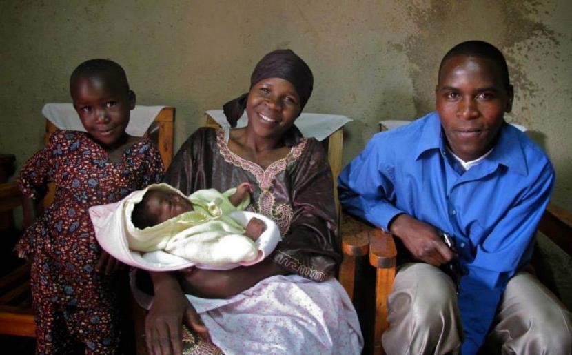 A smiling family sits together indoors. A woman wearing a headscarf holds a newborn wrapped in a light-colored blanket. A man in a blue shirt sits beside her. A young child stands next to the woman, dressed in a colorful outfit. Wooden furniture is in the background.