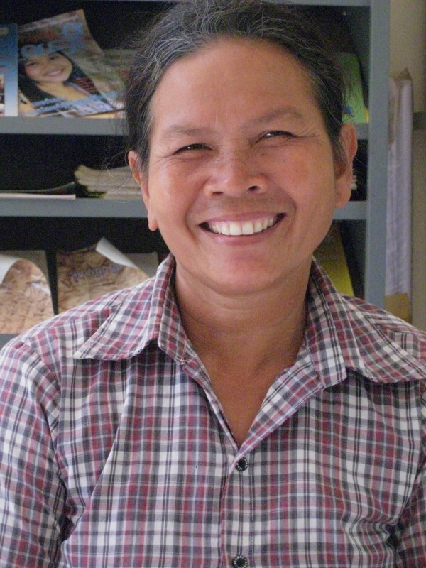 A smiling person with short dark hair and a gray checkered shirt sits indoors. Shelves in the background have books and various papers.