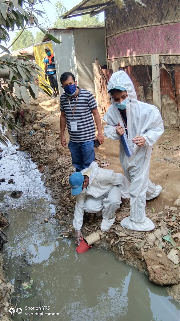 Two people in protective suits and masks are collecting water samples from a polluted stream with containers, while a third person in a blue shirt and jeans observes. The area around them appears rundown, with makeshift structures in the background.