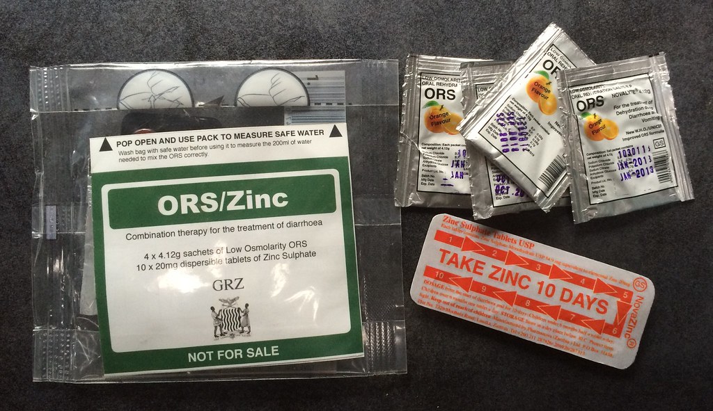 Packets of ORS/Zinc medication are displayed on a table. The package includes instructions for treating diarrhea, sachets of low-osmolarity ORS, zinc tablets for a 10-day course, and a tool for measuring safe water.