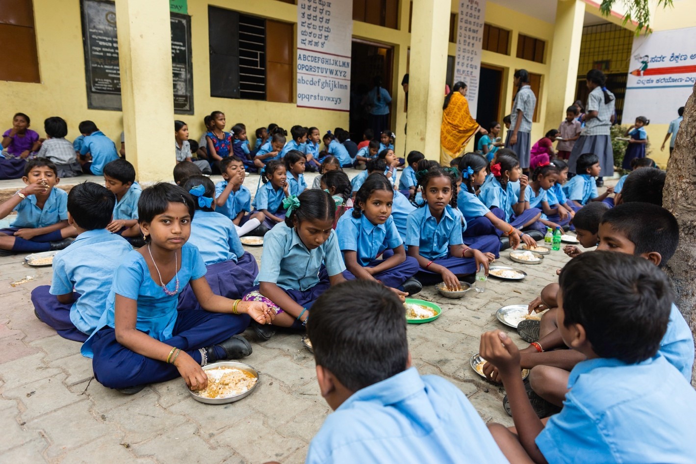 A large group of children in blue uniforms sit on the ground outside a school building, eating meals served in metal plates. The background shows walls with educational posters and additional children and adults standing or sitting.