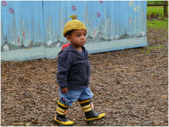 A young child wearing a knitted hat, a dark hoodie, and striped rain boots walks on a wood chip path. Behind them is a blue fence with murals of flowers. The child appears to be outdoors in a garden or park area.