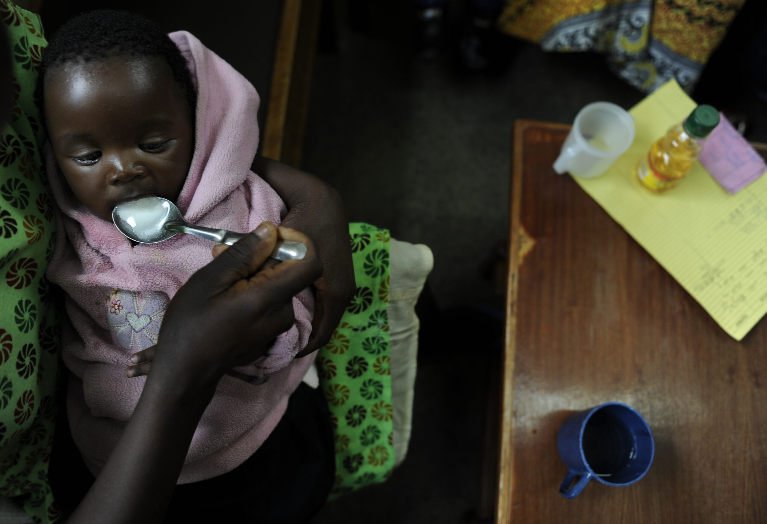 A baby wrapped in a pink blanket is being fed with a spoon by an adult's hand. The scene is set indoors, with a table nearby holding a pen, paper, a bottle, and a cup. The baby looks calm and focused on the spoon.