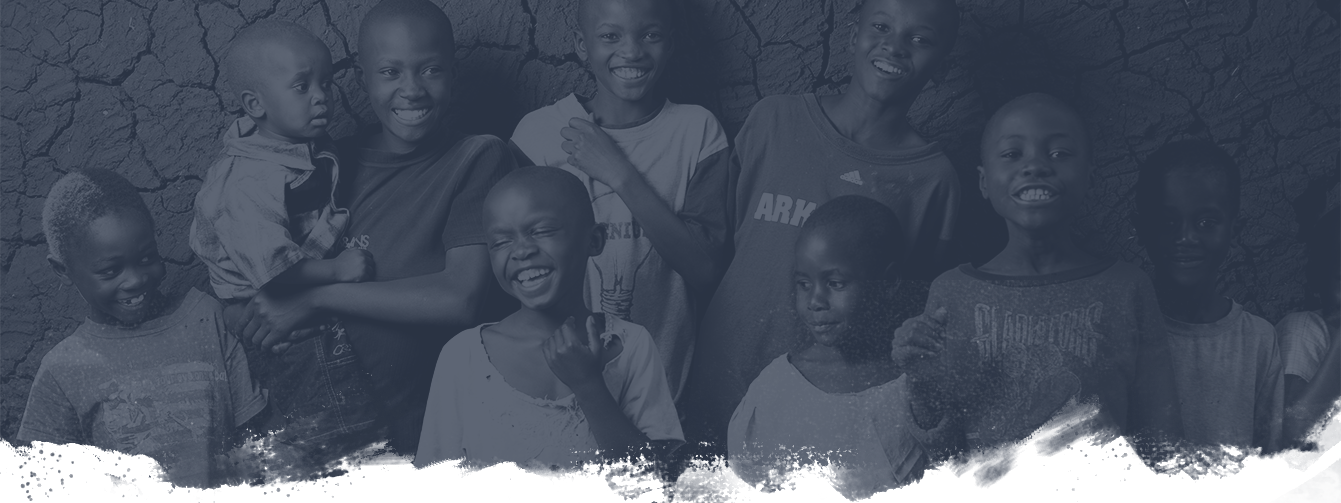 A group of smiling children stand in front of a textured, mud wall. The children are dressed in casual clothes, with some wearing T-shirts and one child holding a toddler. They appear joyful and close-knit.