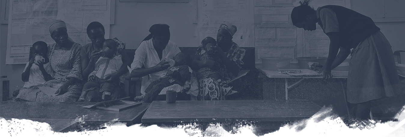 A group of women sit on benches in a waiting area, each holding a young child. The background features several posted notices on the wall. A healthcare worker bends over a table on the right side of the image.