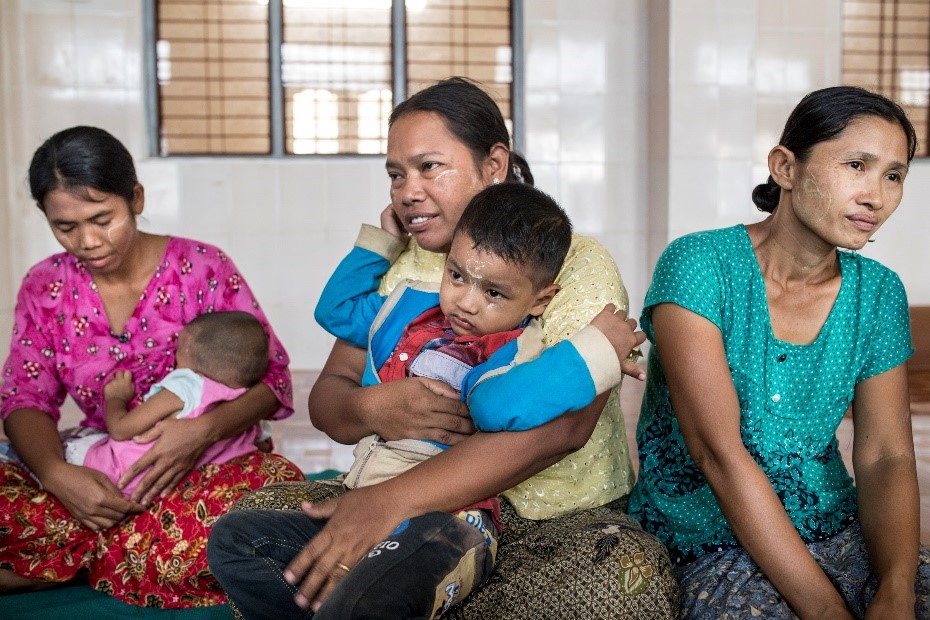 Four women sit closely together indoors with two holding young children. The woman in the center wears a yellow shirt and holds a young boy dressed in red. The women on each side are also caring for a younger child. They appear to be in a casual, communal setting.