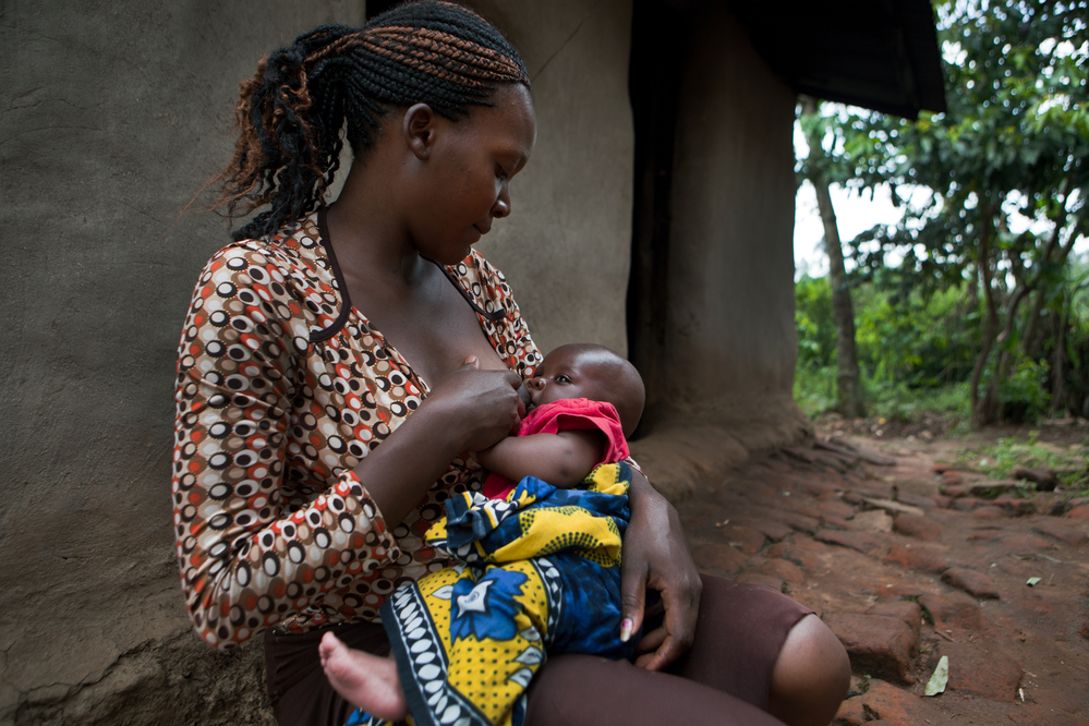 A woman wearing a patterned blouse is sitting outside on a brick path near a mud wall, breastfeeding her baby. The baby is wrapped in a colorful cloth. There is greenery in the background.