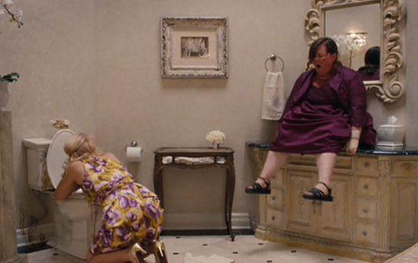 A woman in a floral dress kneels over a toilet, appearing unwell, while another woman in a purple dress sits on a counter, also appearing distressed. The bathroom is elegantly decorated with a large mirror and artwork on the wall.