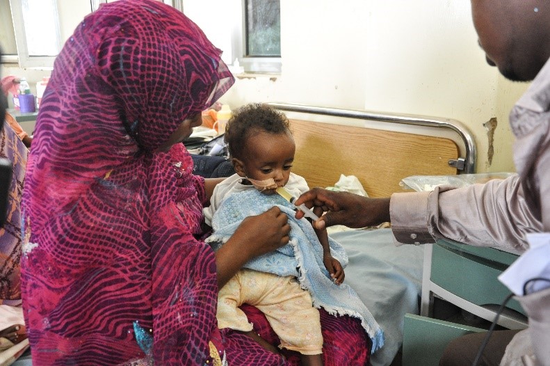 A caregiver dressed in a bright, patterned outfit holds a young child on their lap in a hospital room. The child is being fed or given medicine by a healthcare professional. The room has a bed and medical equipment in the background.