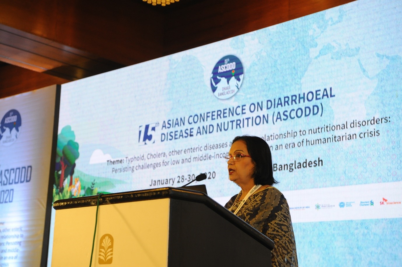 A woman stands at a podium giving a speech at the 15th Asian Conference on Diarrhoeal Disease and Nutrition (ASCODD). The event backdrop behind her carries the conference's details, including the theme focused on persisting challenges for disease control in humanitarian crises.