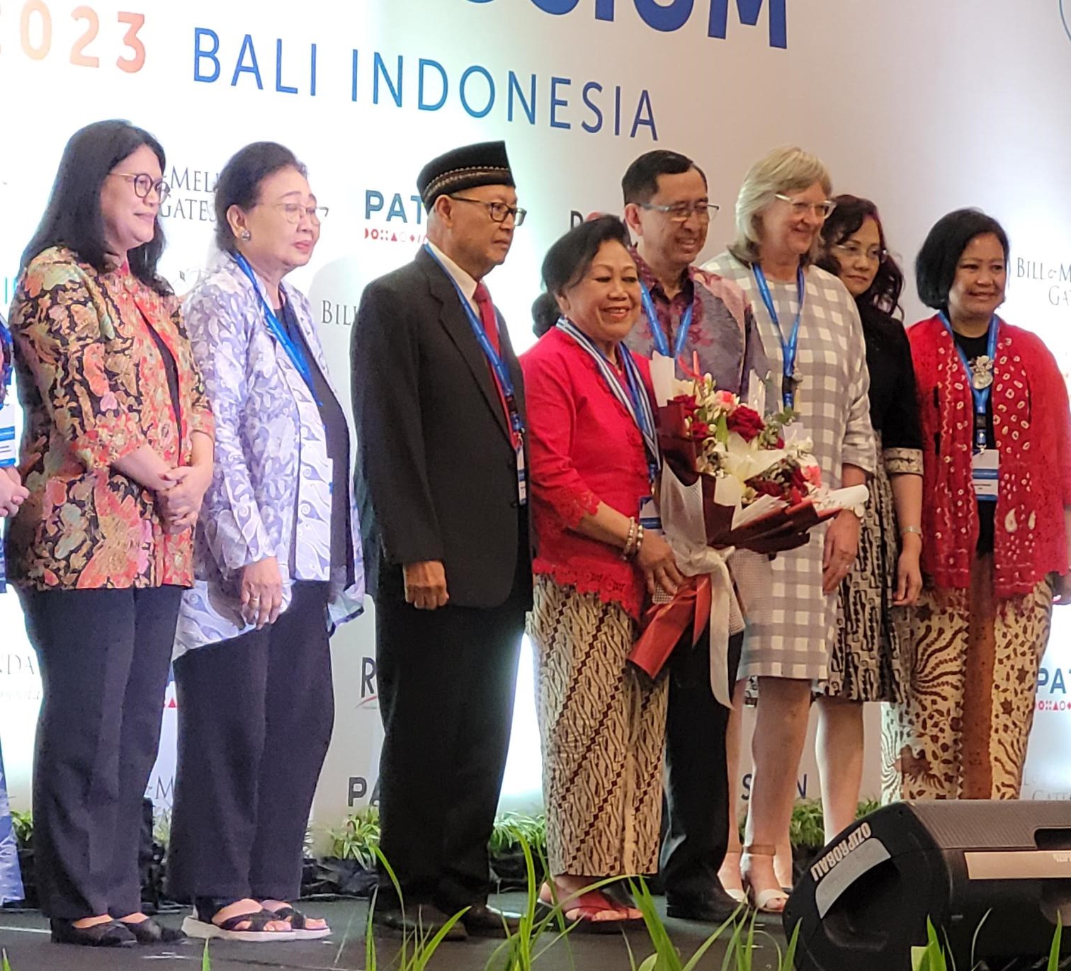 A diverse group of people, wearing formal attire and traditional Indonesian clothing, stand together on a stage. A woman in the center holds a bouquet. The backdrop reads "2023 Bali Indonesia.