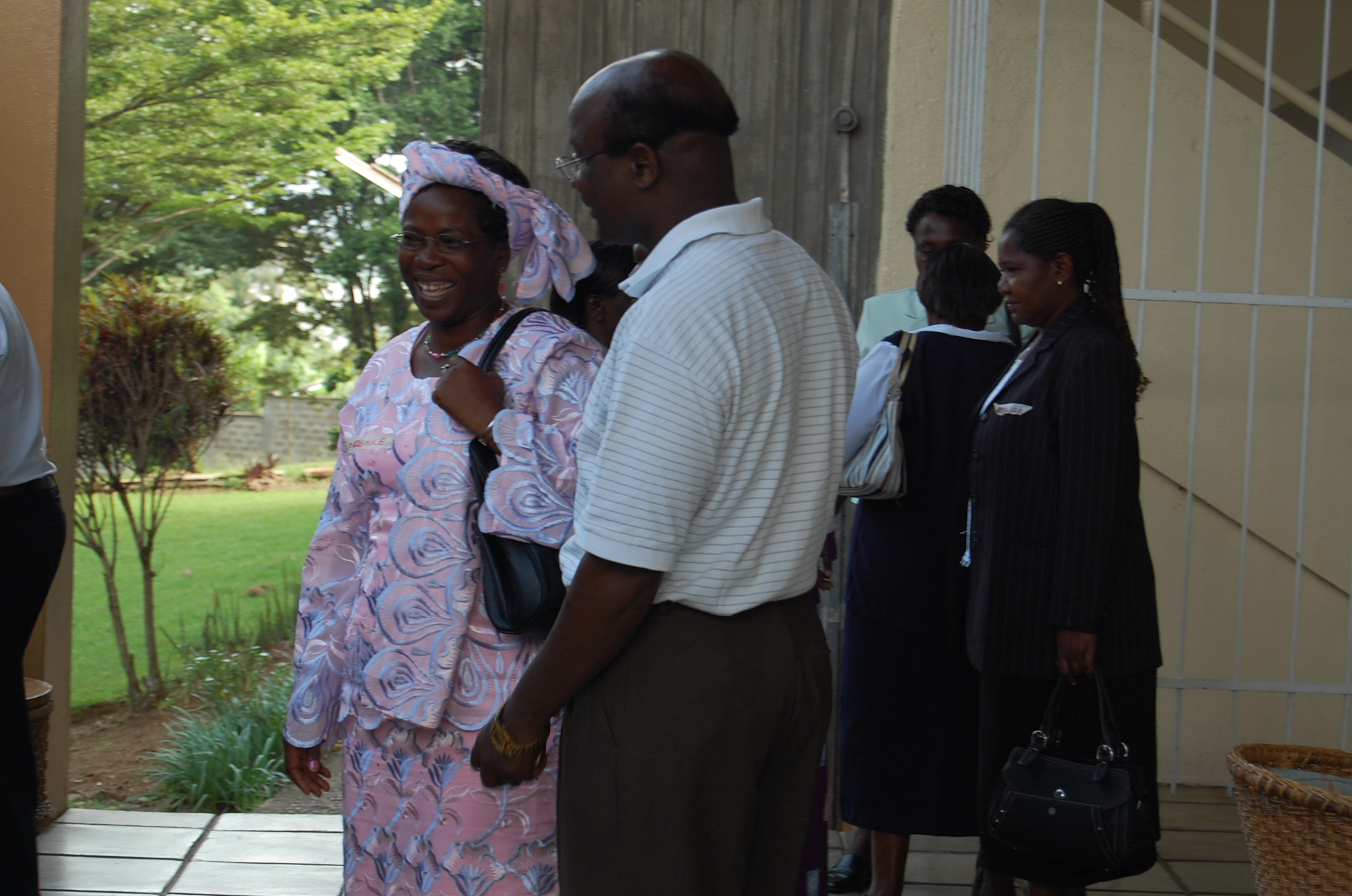 A group of people standing and talking outside a building. A woman in a pink and purple traditional outfit and headscarf is smiling and speaking with a man in a white shirt and brown pants. Additional people are in the background near a gated entrance.