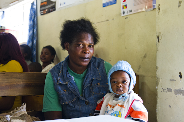 A woman wearing a denim vest and green shirt sits on a wooden bench holding a baby dressed in warm clothing and a blue hat. Other people are seated in the background against a wall with posters. The setting appears to be indoors.