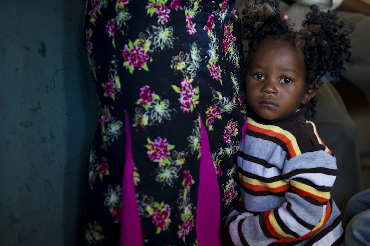 A young child with curly hair and a striped sweater stands next to an adult wearing a dark floral-patterned skirt. The child looks at the camera with a calm expression, while holding onto the adult's leg.