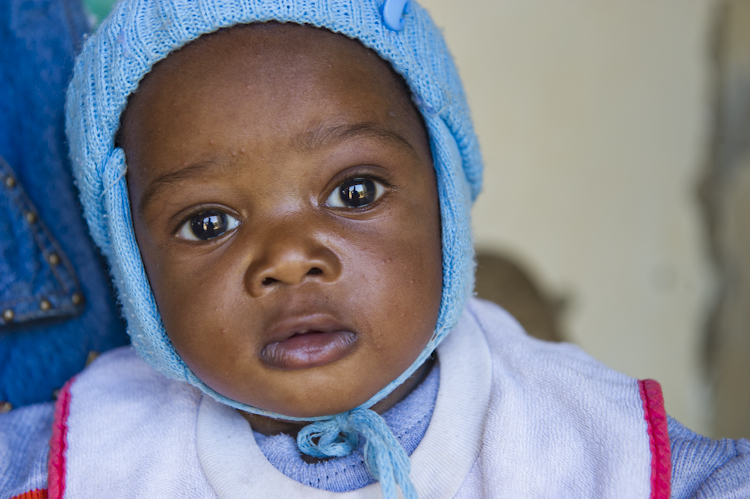 A close-up of a baby wearing a blue knit cap with a string tied under the chin. The baby has a curious expression, with wide eyes and slightly parted lips, and a white and pink outfit. The background is softly blurred.