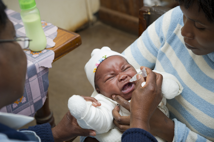 A baby wearing a white outfit and hat is being held by a caregiver while a healthcare worker administers oral medicine or vaccine. The baby appears to be crying. A bottle and a table with a cloth are visible in the background.