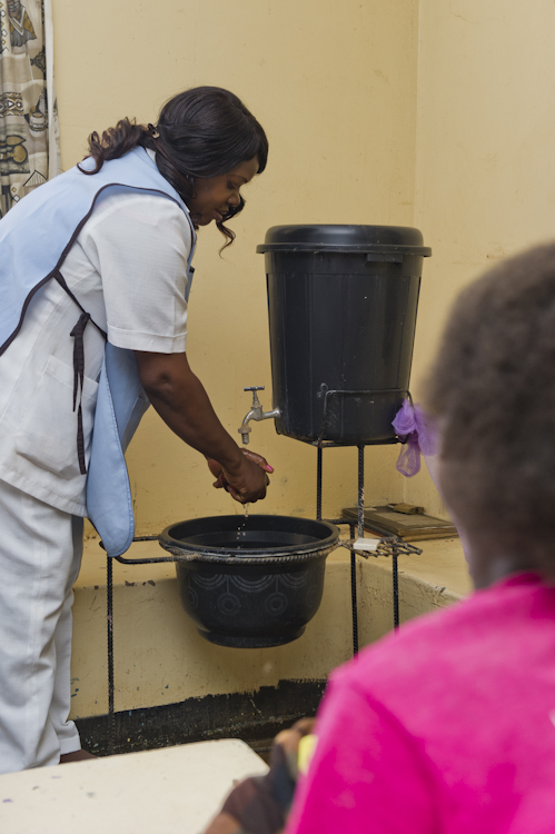 A healthcare worker in a white uniform and blue vest washes her hands at a makeshift sink with a black plastic water dispenser. The wall behind her is yellow, and a person in a red shirt is in the foreground, slightly out of focus.