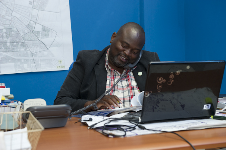 A man sits at a cluttered desk in an office, smiling as he talks on a corded phone. He is writing or reviewing documents. In front of him is a laptop and behind him is a wall with a large map.