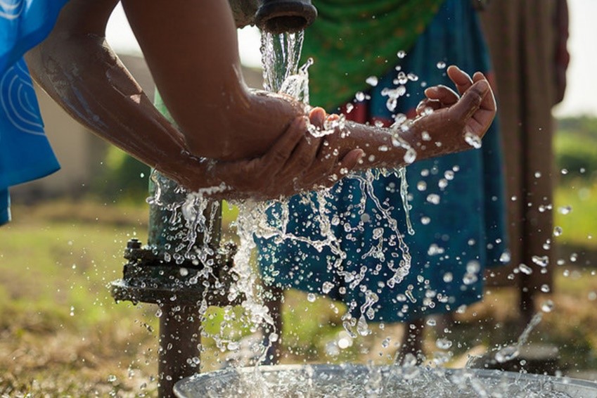 A close-up of hands being washed under a flowing outdoor faucet, with water splashing in all directions. The background shows blurred figures and greenery, indicating an outdoor rural setting.