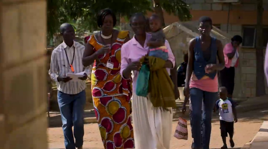 Several adults and children walk outdoors near a building. One woman, dressed in a colorful dress, walks while holding a child in her arms. The group appears to be in a sunny, open environment with trees casting shadows on the ground.