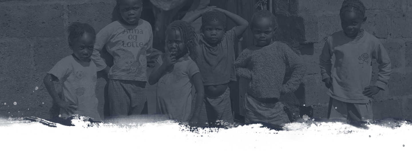 A group of six children stands in front of a brick wall. They are looking at the camera with varying expressions. Some children have their hands on their hips, while one has her hand above her head. All are dressed in casual clothing. The image is monochromatic.