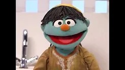 A blue puppet with shaggy black hair, a round orange nose, and wide eyes is smiling. The puppet is dressed in a beige shirt with decorative patterns and standing in front of a white background with a blurry faucet in the background.