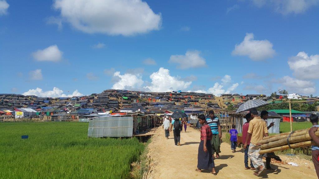 People walk along a dirt path surrounded by green fields and makeshift shelters under a bright blue sky with scattered clouds. The shelters extend up a hillside in the background, indicating a densely populated area. Several individuals carry bamboo poles.