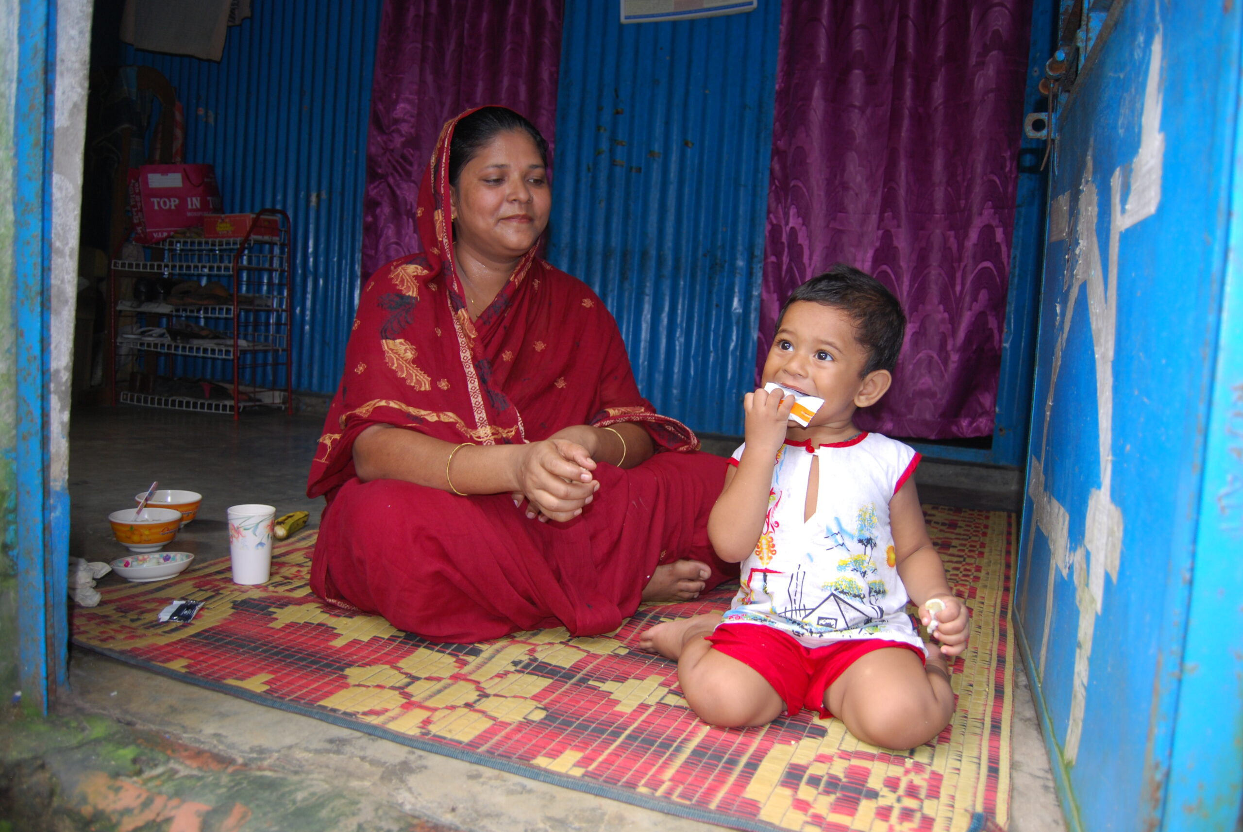 A woman in a red sari sits on a colorful mat in a small room next to a child wearing a white top and red shorts. The child is eating, and various food items are placed on the floor near them. The room has blue walls and purple curtains.