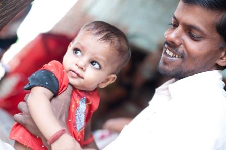 A smiling man gently holds a baby in his arms. The baby is wearing a red shirt and looking curiously to the side. The background is softly blurred, suggesting a cozy, indoor setting.