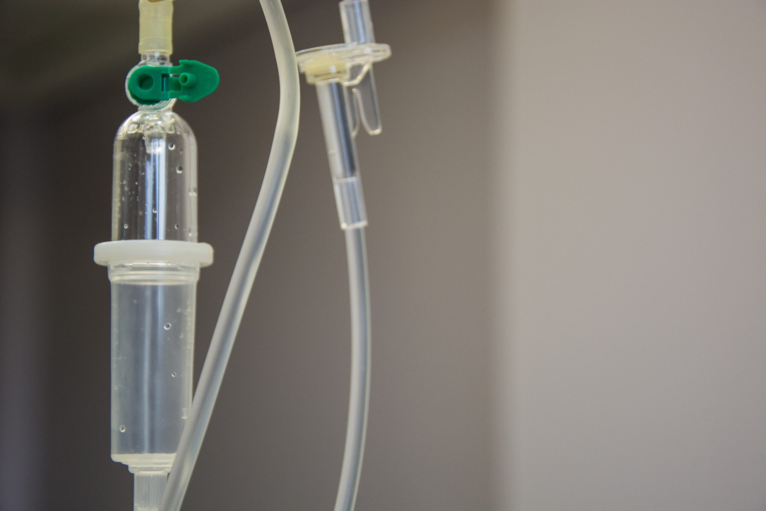 Close-up of a medical IV drip set against a neutral, blurred background. The drip chamber and tubing are visible, with a green clamp on one tube.