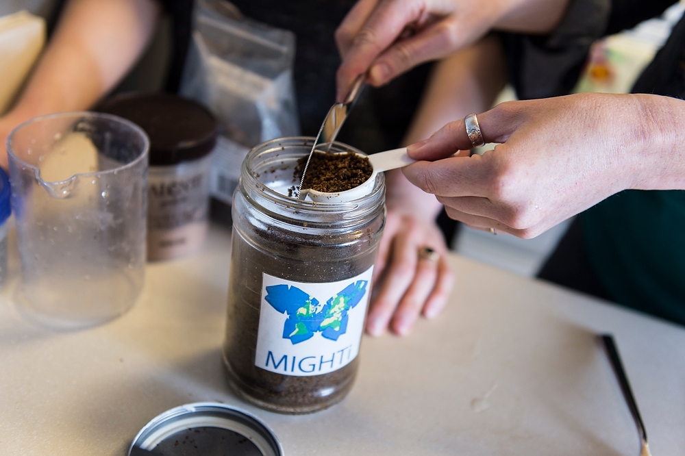 Two people measuring coffee grounds from a jar labeled "Mighty" into a scoop. A beaker and other containers are on the table. One person is wearing a ring on their left hand.