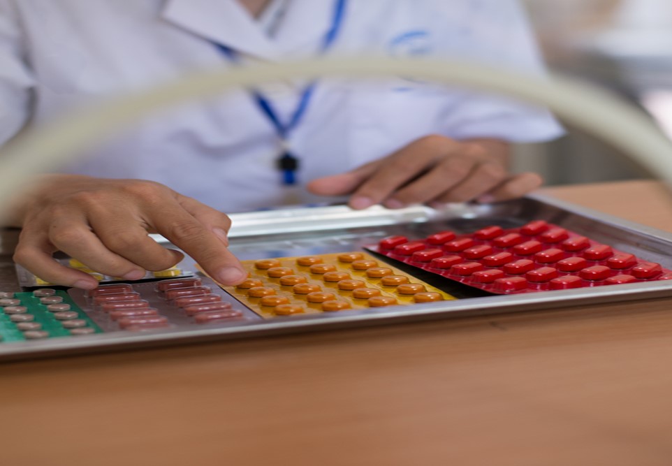 A person in a lab coat is sorting colorful blister packs of pills on a tray. There are green, yellow, orange, and red tablets organized in rows. The person's face is not visible, only their hands and part of their torso.