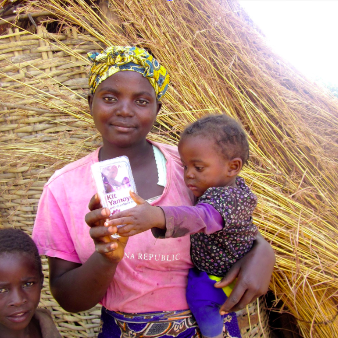 A woman stands holding a small child on her hip. She wears a colorful headscarf and a pink shirt while smiling at the camera, holding a packaged item labeled "Kit Yamoyo." A young boy stands beside them. Behind them is a woven structure made of straw.