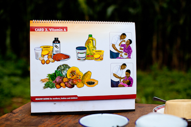 A visual guide titled "CARD 3. Vitamin A" displays food items rich in Vitamin A, including eggs, fish, liver, various vegetables and milk. Beside it are three diagrams showing a person feeding a child. The guide is placed on a wooden surface outdoors.