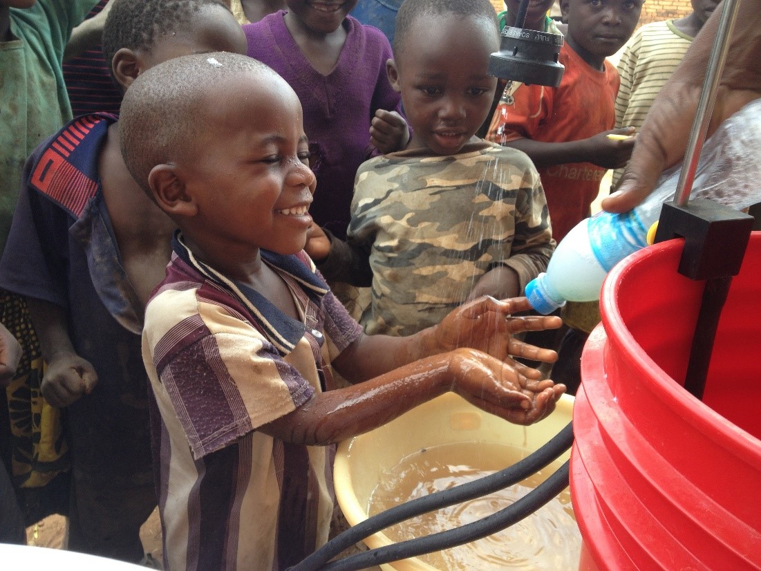 Children smile and excitedly wash their hands with soapy water near a bright red container outdoors. Two kids, one wearing a striped shirt and another in a camo shirt, look on while an adult helps pour water from a bottle. Other children observe in the background.