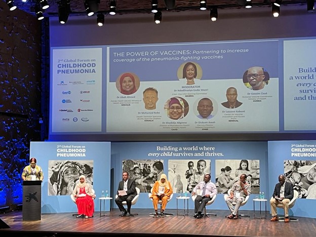 A panel discussion at the 2nd Global Forum on Childhood Pneumonia, featuring six speakers seated on stage. A large screen behind displays the topic "THE POWER OF VACCINES: Partnering to increase coverage of the pneumonia-fighting vaccines.