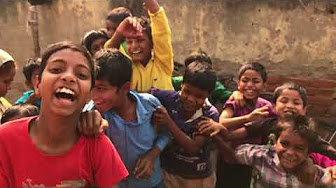 A group of children, beaming with wide smiles and laughter, stand closely together against a weathered, outdoor backdrop. They appear to be in a joyful mood, with some raising their hands and others playfully embracing each other.