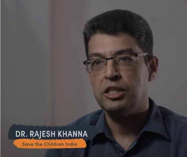 A man with glasses and dark hair, identified by the text on the lower left as "Dr. Rajesh Khanna, Save the Children India," is speaking in a video. The background is blurred, focusing attention on him.