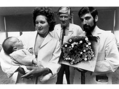 A woman holds a baby wrapped in a blanket, while two men stand beside her. One man holds a scientific image of smallpox virus particles. All are dressed in lab coats, suggesting a medical or scientific setting.