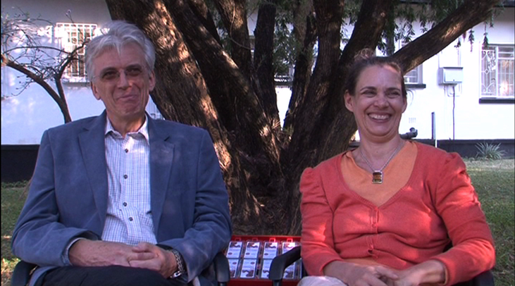 A man and a woman are sitting outdoors on a bench in front of a tree and smiling. The man, wearing a blue blazer and white shirt, has gray hair. The woman, wearing an orange cardigan, has brown hair pulled back. Both appear relaxed and happy.