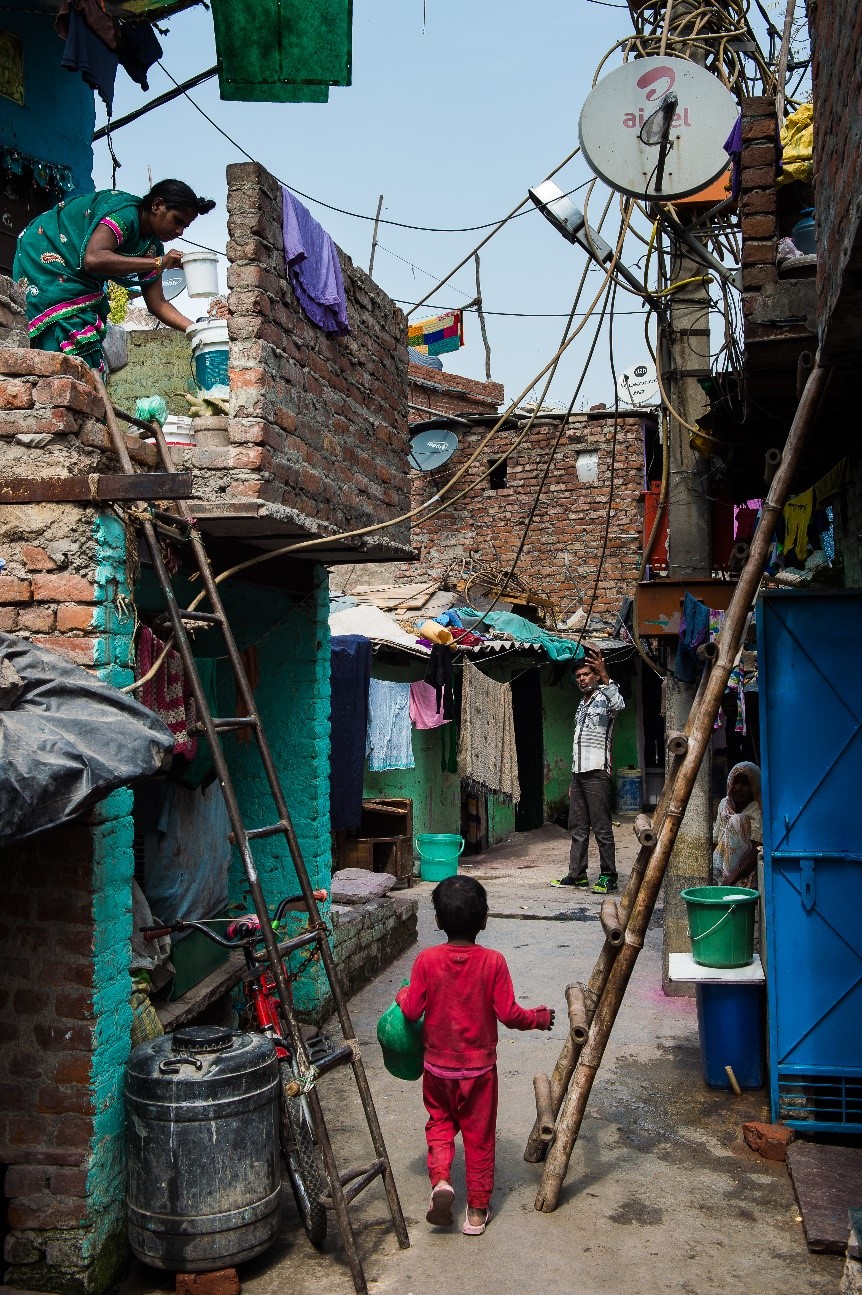 A child in red clothing walks through a narrow, colorful alley of a brick-built neighborhood, holding a green bowl. A woman on the left does laundry, standing on a ladder. A man in the background gestures while standing. Various hanging laundry and cables are visible.