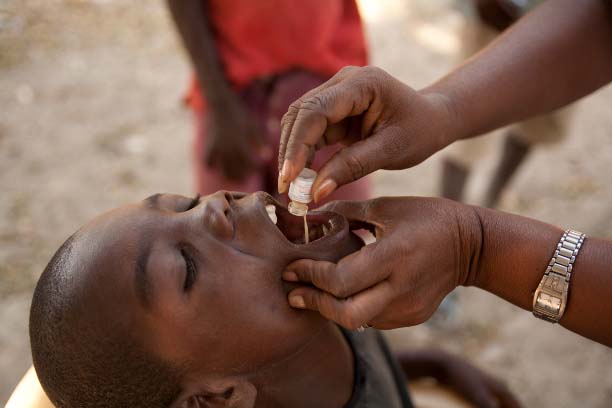 A child receives oral medication from an adult's hands, with the child tilting their head back. The adult holds a small bottle and administers drops into the child's mouth. The scene takes place outdoors with blurred people in the background.