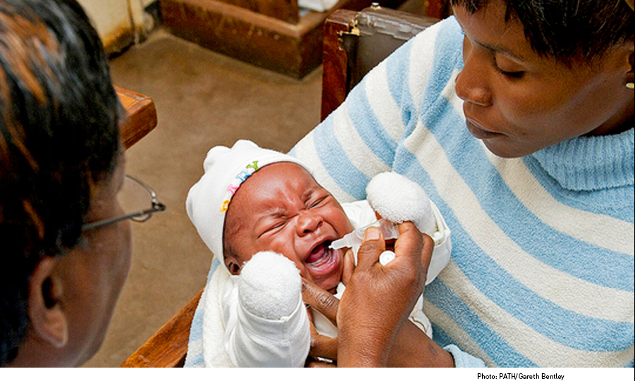 A crying infant in white clothing and a hat is being held by an adult. Another adult is administering oral drops to the baby using a dropper. The scene appears to be in a healthcare setting.