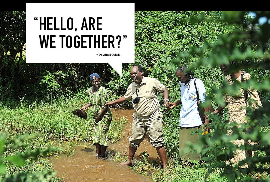 Three individuals are crossing a shallow stream in a forested area, holding hands for support. One person is seen looking and smiling at the camera. A caption superimposed on the image reads, "Hello, are we together?" attributed to Dr. Alfred Odolia.