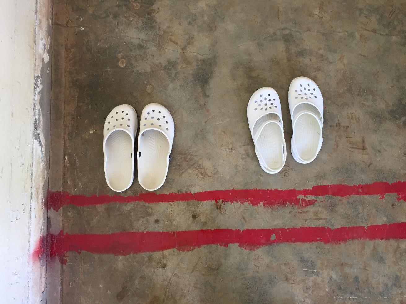 Two pairs of white slip-on shoes are placed on a concrete floor with two parallel red lines. The shoes are positioned on either side of the lines, suggesting separation or division. The floor has a slightly worn texture.