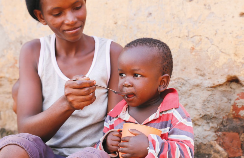 A woman smiles as she feeds a young child with a spoon. The child, sitting on her lap and holding an orange cup, looks content while being fed. They are outside, against a rustic wall, in casual attire.