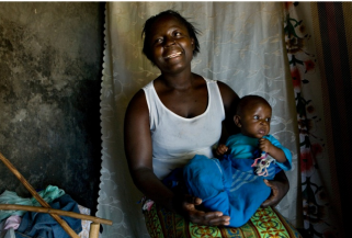 A woman with a wide smile is sitting indoors, holding a baby wrapped in a blue blanket. She is wearing a white tank top and patterned skirt. The background features a white curtain and some household items. Both the woman and baby appear happy and content.