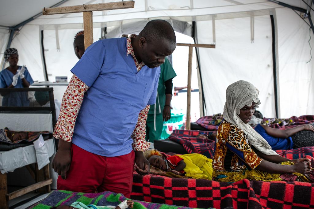 A healthcare worker in blue scrubs tends to patients lying on beds under a white tent. The patients are covered with colorful blankets, and another healthcare worker is seen in the background. The setting appears to be a temporary medical facility.