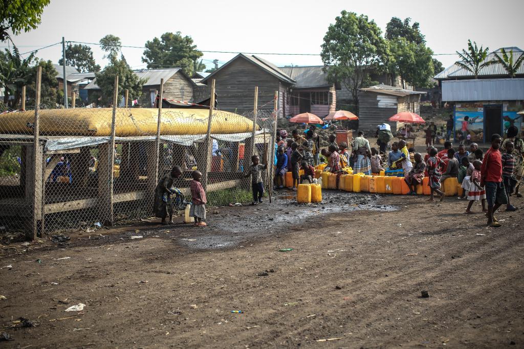 A large group of people, many of them children, gather with yellow containers near a water storage area. They appear to be collecting water. The scene is set in a community with modest houses and some trees in the background. The ground is muddy.